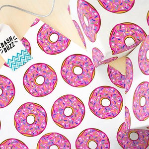 K&ouml;nnte beinhalten: Wei&szlig;es Geschenkpapier mit einem sich wiederholenden Muster aus rosafarbenen Donuts mit bunten Streuseln. Die Donuts haben eine wei&szlig;e Mitte und einen gelben Umriss. Der Text "Bash Buzz" befindet sich auf einem t&uuml;rkisfarbenen und wei&szlig;en Etikett.