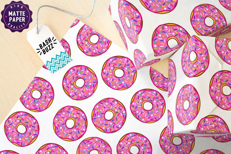 K&ouml;nnte beinhalten: Wei&szlig;es Geschenkpapier mit einem sich wiederholenden Muster aus rosafarbenen Donuts mit bunten Streuseln. Die Donuts haben eine wei&szlig;e Mitte und einen gelben Umriss. Der Text "Bash Buzz" befindet sich auf einem t&uuml;rkisfarbenen und wei&szlig;en Etikett.