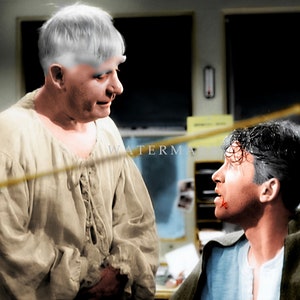 May include: Two men in a scene from a movie or play. The man on the left is wearing a light brown shirt and has white hair. The man on the right is wearing a blue shirt and has dark hair. He has a small cut on his face.