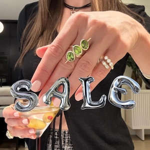 Puede incluir: Un anillo de oro con tres piedras preciosas de peridoto verde de corte ovalado dispuestas en fila, usado junto a otro anillo de oro con varias piedras claras redondas pequeñas. La palabra SALE aparece en grandes letras plateadas metálicas.