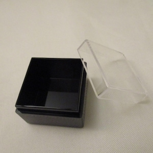 Puede incluir: Una caja cuadrada negra con una tapa de plástico transparente. La caja está vacía y la tapa está abierta.