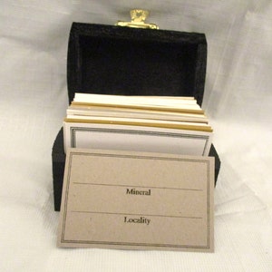 May include: A small, black wooden chest with a gold-colored clasp, filled with index cards. One card is in front, with the words "Mineral" and "Locality" printed on it. The cards have a gold border.