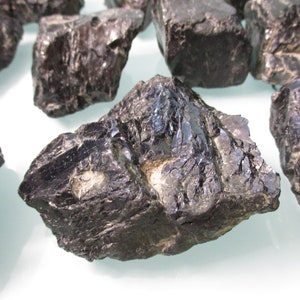 May include: A collection of rough, dark black stones with a glossy, reflective surface. The stones have irregular shapes and textures, with visible cracks and facets. The lighting highlights the natural sheen of the mineral specimens.