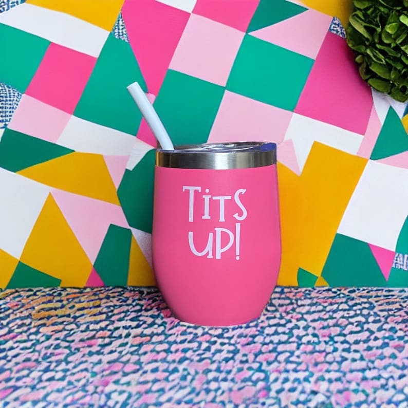 May include: A pink insulated tumbler with a silver rim and a white straw. The tumbler has the words "Tits Up!" in white lettering. The background features a colorful geometric pattern with pink, green, yellow, and white shapes.
