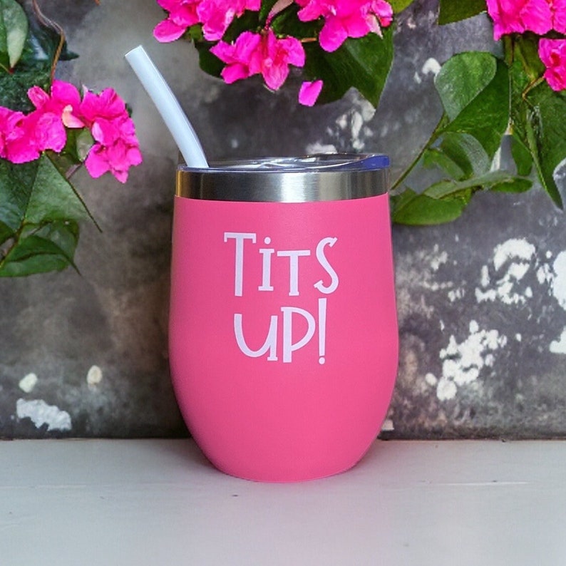 May include: A pink insulated tumbler with a silver rim and a white straw. The tumbler has the words "Tits UP!" printed in white. The background includes pink flowers and a gray wall.