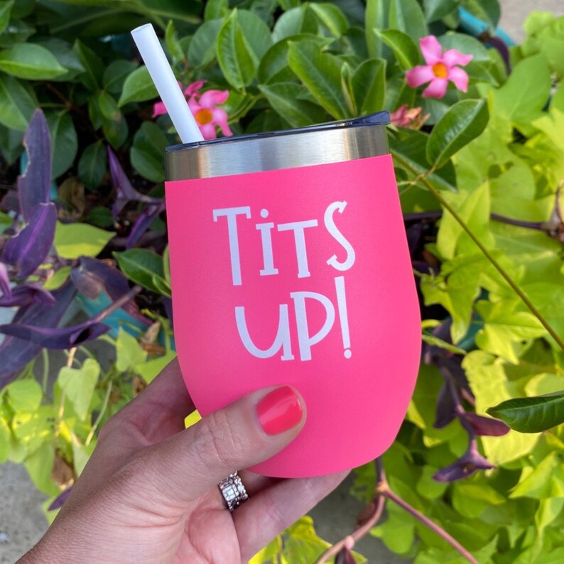 May include: A bright pink stemless wine tumbler with the words "Tits UP!" in white. The tumbler has a silver rim and a white straw. The tumbler is held in front of a background of green and purple foliage.