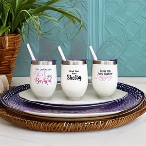Puede incluir: Tres vasos de vino de acero inoxidable blanco con tapas y pajitas. Los vasos tienen diferentes lemas impresos: "Mis colores son Blush y Bashful", "Bebe tu zumo Shelby", y "Te quiero m&aacute;s que a mi equipaje".
