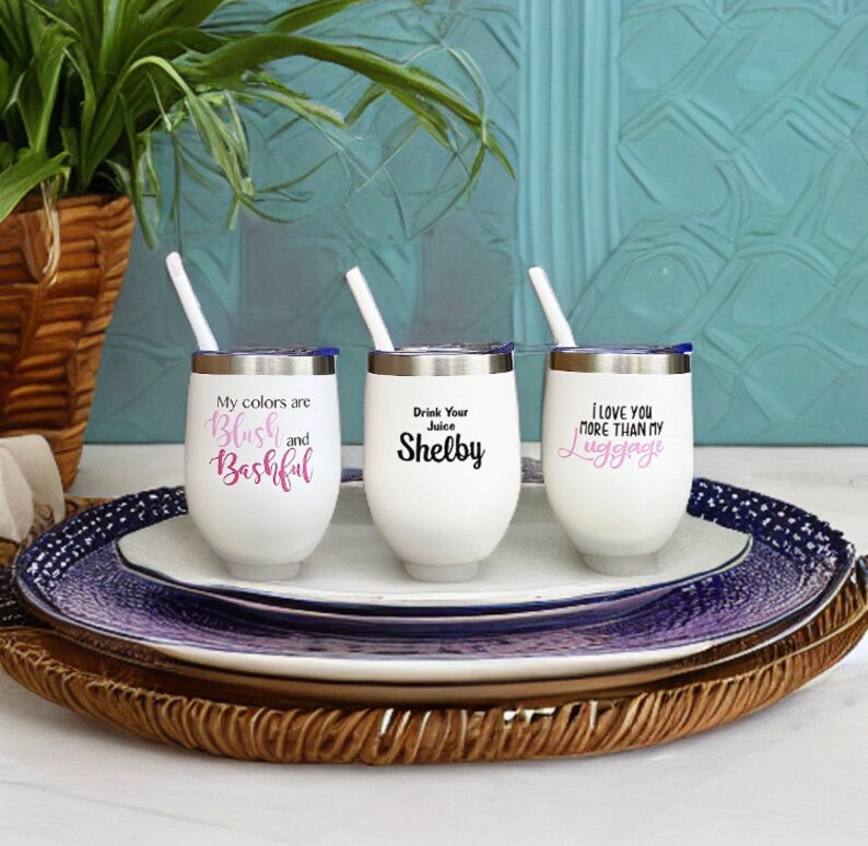 Puede incluir: Tres vasos de vino de acero inoxidable blanco con tapas y pajitas. Los vasos tienen diferentes lemas impresos: "Mis colores son Blush y Bashful", "Bebe tu zumo Shelby", y "Te quiero m&aacute;s que a mi equipaje".