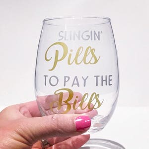 May include: A clear glass wine glass with a gold vinyl decal that reads "Slingin' Pills to Pay the Bills".
