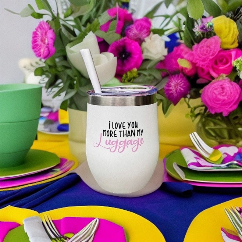 Puede incluir: Un vaso t&eacute;rmico de acero inoxidable blanco con una tapa rosa y una inscripci&oacute;n en negro y rosa que dice "i love you more than my luggage". El vaso est&aacute; sobre una mesa con un mantel azul y manteles individuales coloridos.