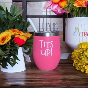 May include: A pink insulated tumbler with a silver rim and a white straw. The tumbler has the words "Tits Up!" printed in white. The tumbler is sitting on a wooden surface, with flowers in the background.