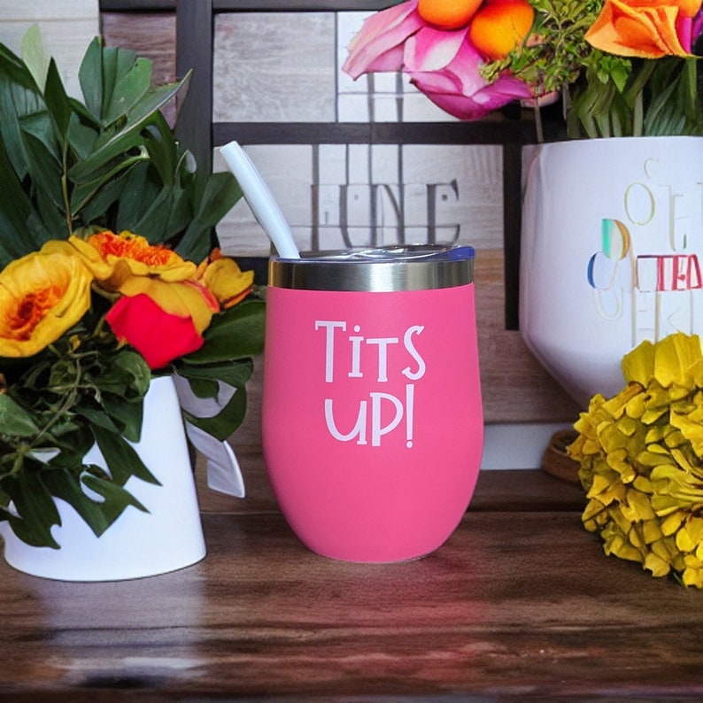 May include: A pink insulated tumbler with a silver rim and a white straw. The tumbler has the words "Tits Up!" printed in white. The tumbler is sitting on a wooden surface, with flowers in the background.
