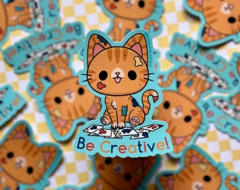 May include: Orange and white cat sticker with paint splatters on its fur. The cat is sitting on a pile of colorful paint splattered paper. The sticker has the text "Be Creative!" on a blue background.