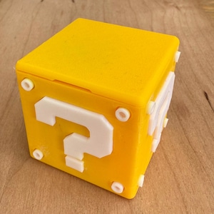 May include: A yellow cube featuring a white question mark, resembling a video game block. The cube has white circular accents on each corner. The block is displayed on a wooden surface.