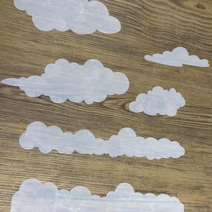 May include: Five white cloud shapes cut from paper, arranged on a woodgrain surface. The clouds are different sizes and shapes, with some having a more rounded appearance and others having a more jagged appearance.