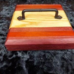 May include: A wooden serving tray with a black metal handle. The tray is made of different types of wood, including light wood, red wood, and dark wood. The tray has a rectangular shape and is approximately 12 inches long and 8 inches wide.