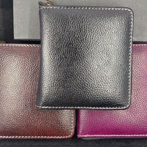May include: Three leather wallets in different colors: black, burgundy, and purple. Each wallet is square-shaped with a zipper closure and white stitching around the edges. The wallets have a textured surface.