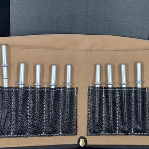 May include: A tan and black leatherette case with a zipper, holding several silver tools. The tools are cylindrical with rounded tops and are arranged in black pockets. A dark green and black tool is secured with a black strap on the right.