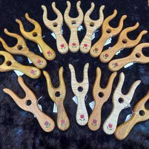 May include: A collection of wooden yarn forks, each with a unique grain pattern and a painted red tulip design. These tools are designed for crafting and are arranged on a dark surface.