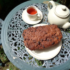 May include: A close-up of a chocolate cake with a cup of tea on a green and white patterned table. The cake is on a white plate with a gold rim.