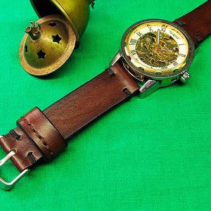 May include: A close-up of a mechanical watch with a brown leather strap. The watch face has a gold-colored skeleton design with Roman numerals. A small, antique brass container with star-shaped cutouts is in the background.
