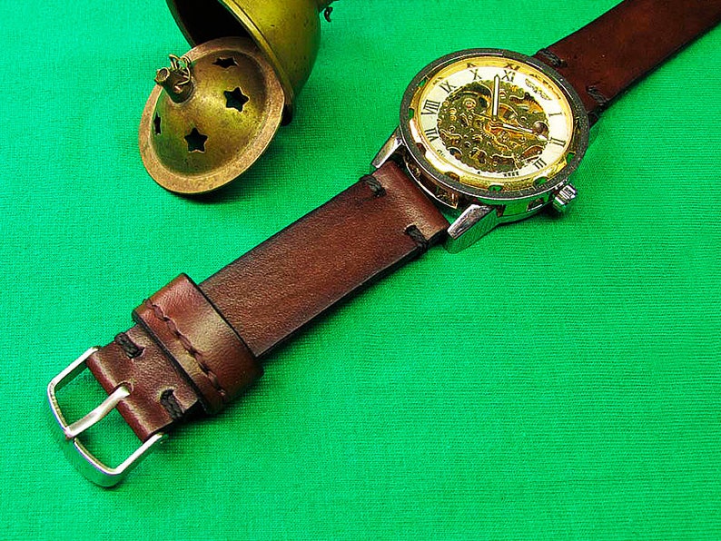 May include: A close-up of a mechanical watch with a brown leather strap. The watch face has a gold-colored skeleton design with Roman numerals. A small, antique brass container with star-shaped cutouts is in the background.