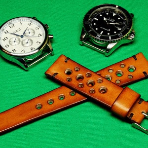 May include: Two wristwatches with brown leather watch bands with holes. The watch bands are crossed over each other. The watch bands have silver buckles. One watch has a white face with black hands and black numbers. The other watch has a black face with silver hands and silver numbers.