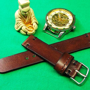 May include: Two brown leather watch straps with silver buckles are crossed on a green surface. A silver and gold skeleton watch and a small ivory-colored figurine are also present. The leather straps have visible stitching.