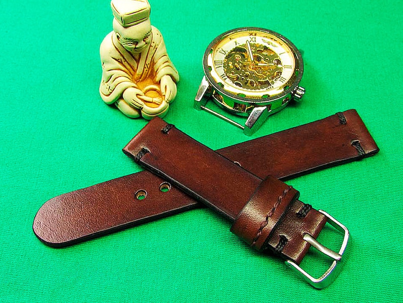 May include: Two brown leather watch straps with silver buckles are crossed on a green surface. A silver and gold skeleton watch and a small ivory-colored figurine are also present. The leather straps have visible stitching.