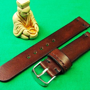 May include: A brown leather watch strap with a silver buckle. The strap has several holes for adjustment and is stitched with black thread. A small, cream-colored figurine of a seated figure is in the background.