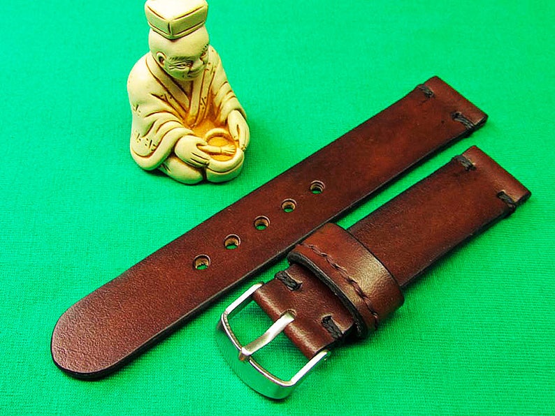 May include: A brown leather watch strap with a silver buckle. The strap has several holes for adjustment and is stitched with black thread. A small, cream-colored figurine of a seated figure is in the background.