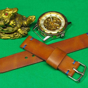 May include: A brown leather watch strap with blue stitching and a silver buckle. The strap is lying on a green surface with a gold turtle figurine and a silver watch with a white face and Roman numeral markings.