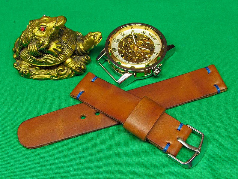 May include: A brown leather watch strap with blue stitching and a silver buckle. The strap is lying on a green surface with a gold turtle figurine and a silver watch with a white face and Roman numeral markings.