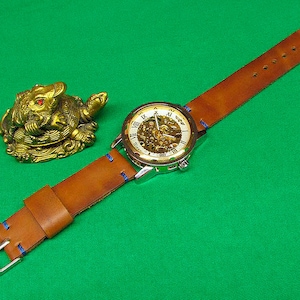 May include: A brown leather watch strap with blue stitching and a silver watch face with Roman numerals. The watch face has a skeleton design, revealing the inner workings of the watch. The watch is on a green surface next to a gold figurine of a turtle.