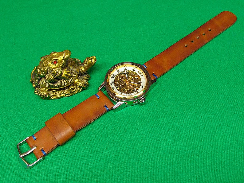 May include: A brown leather watch strap with blue stitching and a silver watch face with Roman numerals. The watch face has a skeleton design, revealing the inner workings of the watch. The watch is on a green surface next to a gold figurine of a turtle.