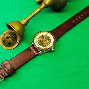 May include: A brown leather watch with a silver case and a skeleton dial, showcasing the inner workings. The watch is paired with a matching brown leather strap and a silver buckle. A brass incense burner is in the background.