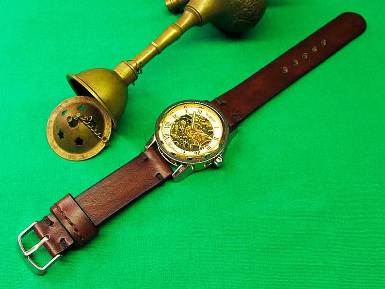 May include: A brown leather watch with a silver case and a skeleton dial, showcasing the inner workings. The watch is paired with a matching brown leather strap and a silver buckle. A brass incense burner is in the background.