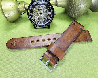 May include: A brown leather watch strap with black stitching. The strap is shown on a green surface with a watch in the background.