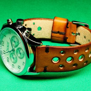 May include: A silver chronograph wristwatch with a white face and brown leather band with holes. The watch has three sub-dials and a tachymeter scale.