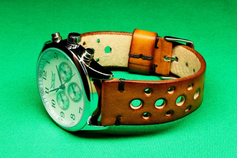 May include: A silver chronograph wristwatch with a white face and brown leather band with holes. The watch has three sub-dials and a tachymeter scale.