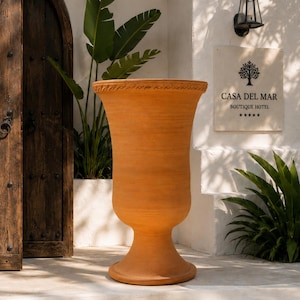 May include: A tall, terracotta-coloured urn with a wide, flared rim featuring a decorative, repeating wave-like pattern. The vessel has a smooth, matte finish, a rounded body that tapers towards a pedestal base, and a wide, circular foot for stability.