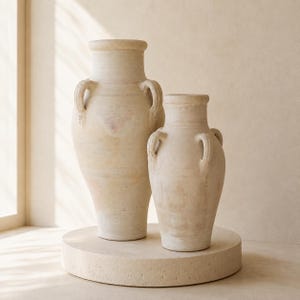 May include: Two off-white ceramic amphora-style vases with a textured, matte finish. Each vessel features a wide neck, a rounded body, and two symmetrical side handles. The vases are displayed in a pair, with one being noticeably taller than the other.