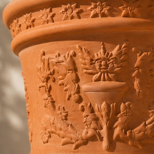 Pode incluir: Um vaso de terracota com um design em relevo de uma urna central ladeada por folhagens e rostos. A borda superior é decorada com um padrão de estrelas repetitivo. O material é argila laranja fosca e terrosa com textura visível.