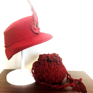 May include: A red hat with a feather and a matching crocheted drawstring bag. The hat is displayed on a white stand. The bag has a tassel and is made of a textured, open-weave material. The items are on a brown surface.