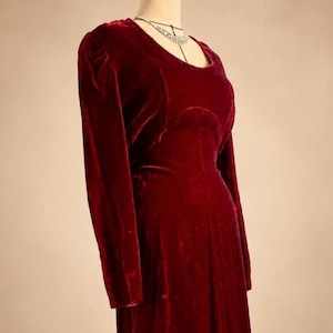 May include: A long-sleeved, deep red velvet dress featuring a rounded neckline and a fitted bodice. The garment displays a smooth, plush texture throughout, with subtle gathering at the shoulders and a clean, structured silhouette.