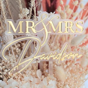 May include: A gold-colored "Mr & Mrs Davidson" cake topper. The topper is made of clear acrylic with gold lettering. The cake topper is surrounded by dried flowers and greenery, with a soft, neutral color palette.