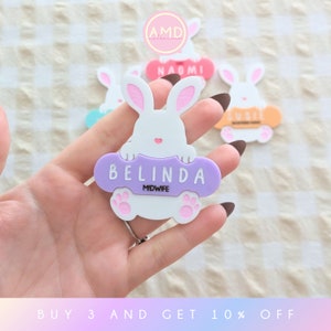 May include: A white bunny holding a purple sign that says "BELINDA MIDWIFE". The bunny is holding the sign with its paws. The sign has a pink paw print on each side.