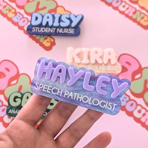 以下が含まれることがあります： 紫と青の大理石模様のネームタグで、名前の「HAYLEY」が紫色の立体文字で、テキストの「SPEECH PATHOLOGIST」が白い文字で書かれています。