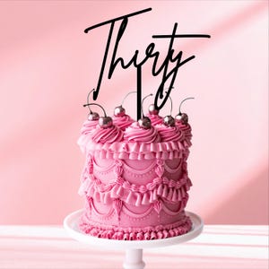 May include: A pink cake with decorative frosting, topped with silver cherries and a black "Thirty" cake topper. The cake sits on a white cake stand against a pink backdrop. A celebratory dessert.