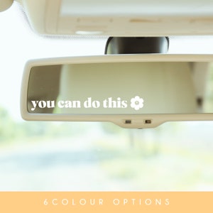 以下が含まれることがあります： 白い車のリアビューミラーに、「you can do this」と白い花柄のアイコンが書かれた白いステッカーが貼られています。